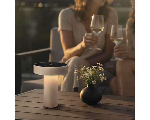 Moderní solární stolní lampa s integrovaným solárním panelem na terasové stole s vázou květin a dvěma osobami držícími sklenice vína.
