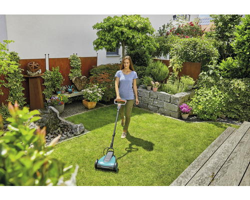 Žena používá sekačku na trávu v udržované zahradě se skalkou a bujnou výsadbou.