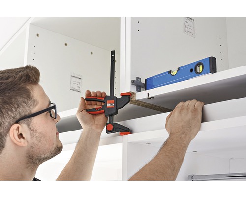 Osoba připevňuje polici svorkou do bílé skříně.