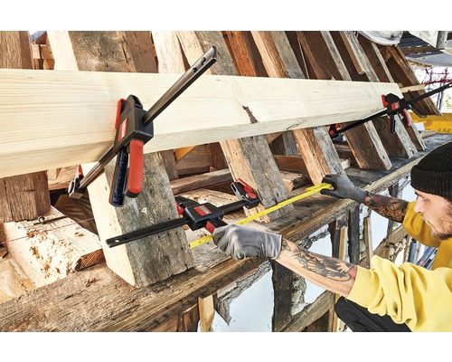 Řemeslník měří kus dřeva měřicím pásmem, které je upevněno šroubovacími svorkami.