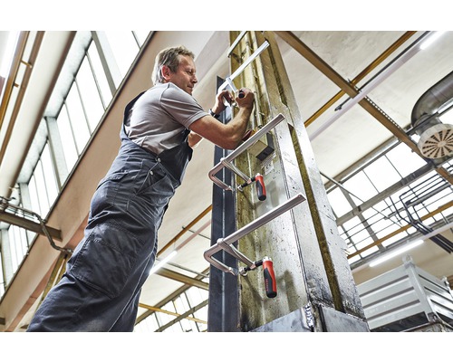 Muž upevňuje dvě šroubovací svorky na kovový nosník v dílně.