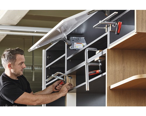 Muž připevňuje čelo nábytku ke skříňce pomocí svorky.