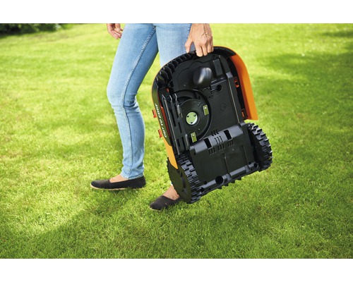 Osoba nese robotickou sekačku přes trávník.