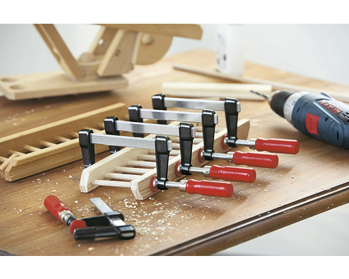 Dřevěný obrobek je upevněn šroubovacími svorkami, vrtačka v pozadí