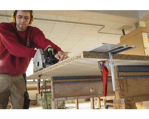 Řemeslník řeže dřevěnou desku ponornou pilou, upevněnou šroubovými svorkami na pracovní stůl