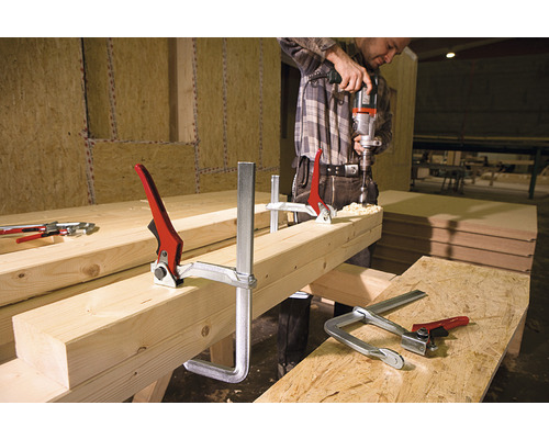 Muž vrtá vrtačkou do kusu dřeva, který je upevněn šroubovacími svorkami.