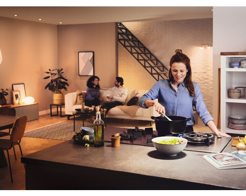 Světlá otevřená kuchyně s ženou vařící u sporáku a lidmi sedícími na pohovce v pozadí.