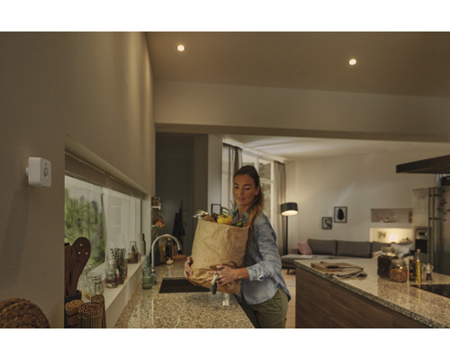 Bílý detektor pohybu na stěně v moderní kuchyni se ženou nesoucí nákupní tašku.