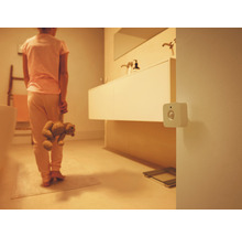 Bílý čtvercový detektor pohybu na stěně v tlumeně osvětlené koupelně s osobou držící plyšového medvídka.