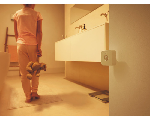 Bílý čtvercový detektor pohybu na stěně v tlumeně osvětlené koupelně s osobou držící plyšového medvídka.