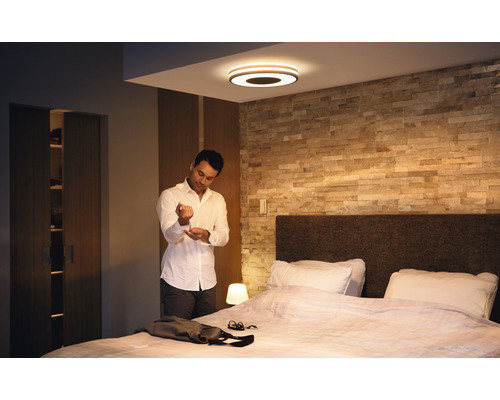 Ložnice se stropním svítidlem, postelí a mužem zapínajícím si košili.