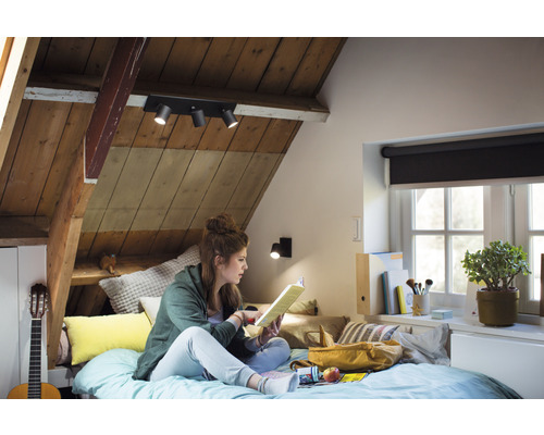 Útulný podkrovní pokoj s dřevěným obložením, vybavený stropním svítidlem s několika reflektory a lampičkou na čtení, kde na posteli sedí a čte mladá žena.