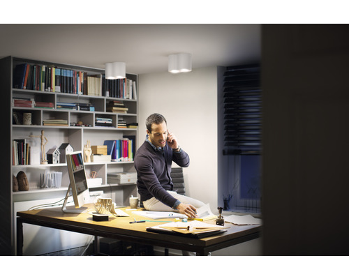 Muž sedí na stole v pracovně s knihovnou a stropními svítidly a telefonuje.