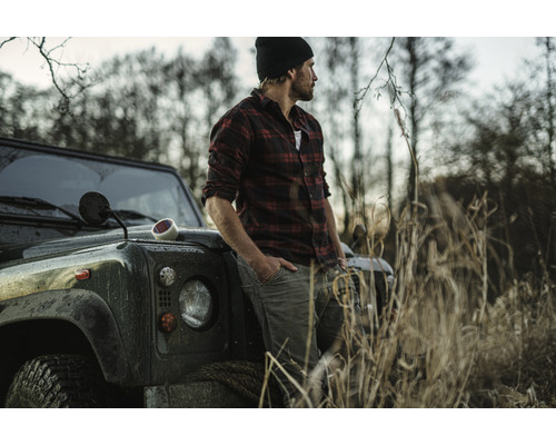 Muž v kostkované košili se opírá o terénní vozidlo ve venkovském prostředí.