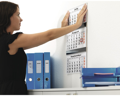 Žena připevňuje kalendář na zeď v kanceláři vedle šanonů a odkládacích košů.