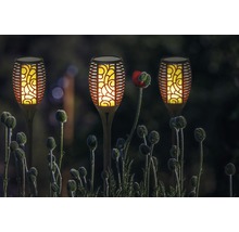 Tři solární lampy ve tvaru pochodní na zahradě mezi kvetoucími rostlinami.