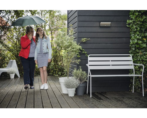 Zahradní scéna se dvěma lidmi pod deštníkem, zahradní lavičkou, rostlinami v květináčích a nástěnným svítidlem na dřevěné stěně.