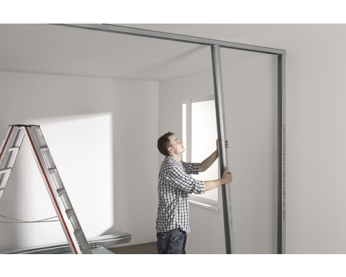 Muž montuje sádrokartonovou stěnu ve světlé místnosti se žebříkem