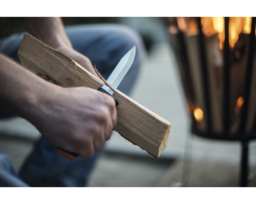 Osoba nožem ořezává dřevo před ohništěm.