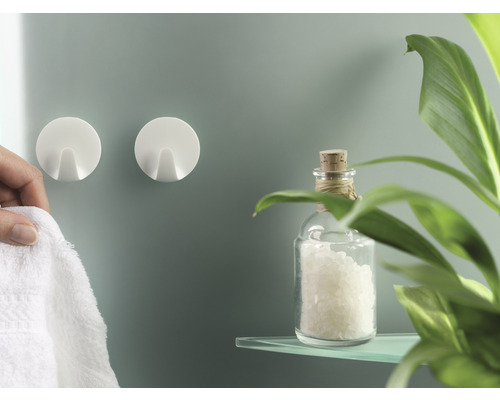 Dva kulaté nástěnné háčky a osoba na ně zavěšuje ručník, spolu se skleněnou lahví a rostlinou na polici