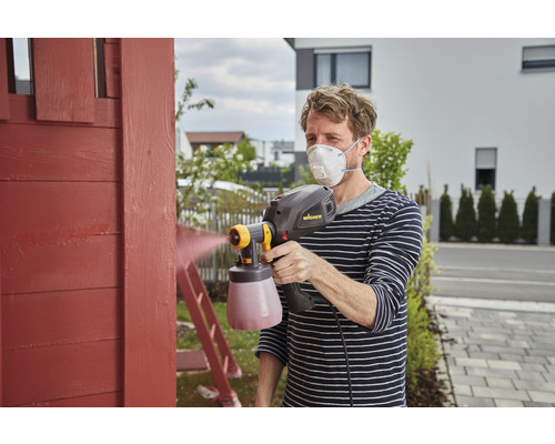 Muž natírá dřevo pomocí stříkacího systému a ochranné masky