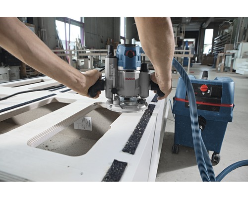 Muž pracuje s horní frézkou na dřevěném obrobku, připojenou k vysavači.