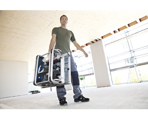 Muž nese stolní kotoučovou pilu v místnosti ve výstavbě.