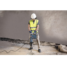 Stavební dělník s ochrannou přilbou, chrániči sluchu, ochrannými brýlemi a výstražnou vestou obsluhuje bourací kladivo k rozbíjení betonu