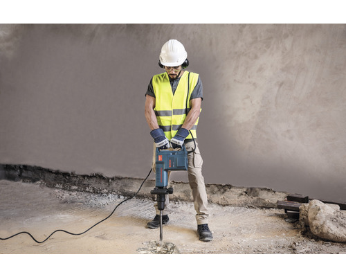 Stavební dělník s ochrannou přilbou, chrániči sluchu, ochrannými brýlemi a výstražnou vestou obsluhuje bourací kladivo k rozbíjení betonu