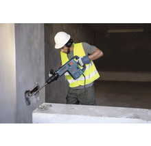 Muž s ochrannou přilbou a bezpečnostní vestou používá bourací kladivo na zeď.