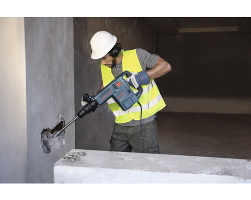 Muž s ochrannou přilbou a bezpečnostní vestou používá bourací kladivo na zeď.