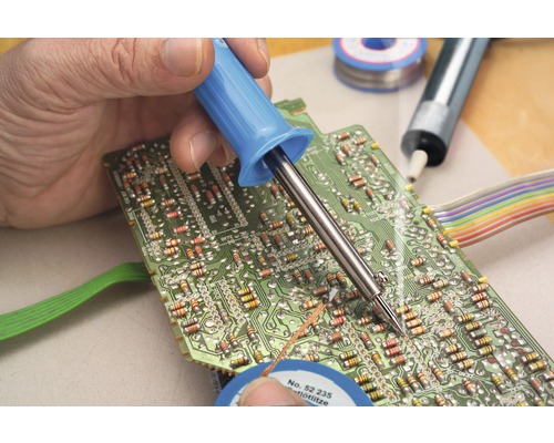 Osoba pájí desku plošných spojů páječkou. K dispozici je také pájka.