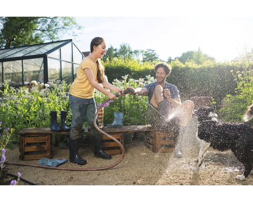 Mladá žena drží zahradní hadici a stříká muže a psa na zahradě. V pozadí je skleník.