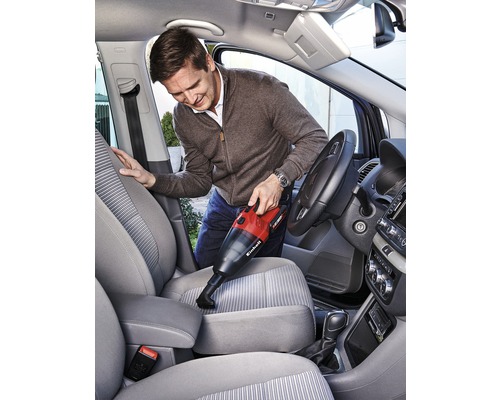 Muž vysává interiér auta pomocí akumulátorového ručního vysavače. Vysavač je červenočerný.