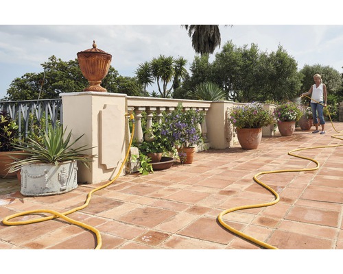 Žena zalévá květiny na terase s terakotovými dlaždicemi a žlutou zahradní hadicí