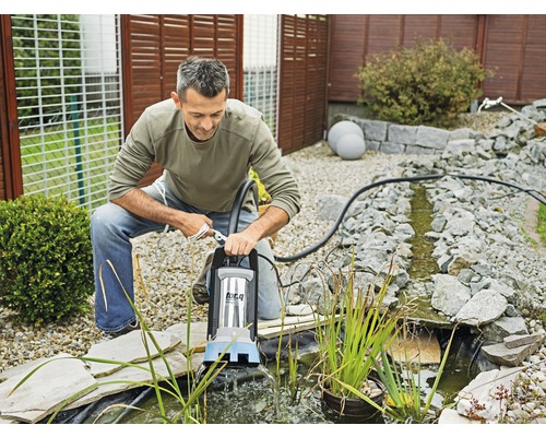 Muž obsluhuje jezírkové čerpadlo na okraji zahradního jezírka, které je obklopeno kameny.