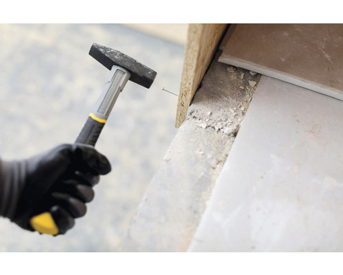 Muž v rukavicích drží kladivo a zatlouká hřebík do dřevěné desky.