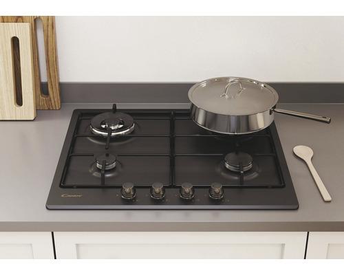 Černý plynový sporák Candy se čtyřmi hořáky, nerezovým hrncem a dřevěnou lžící na pracovní desce.