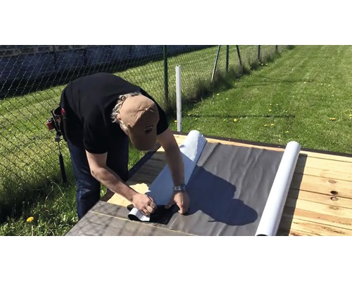 Muž pokládá střešní lepenku na dřevěnou podlahu venku.