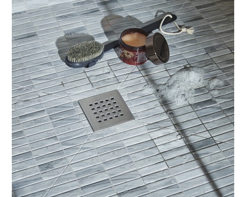 Detailní záběr sprchy s podlahovými dlaždicemi, sprchovým odtokem, kartáčem, přísadou do koupele a mýdlovou pěnou
