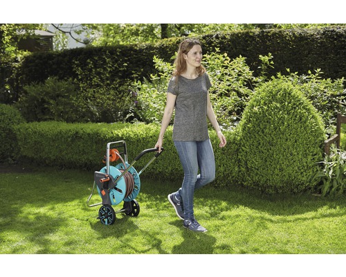 Žena táhne vozík na zahradní hadici po trávníku.