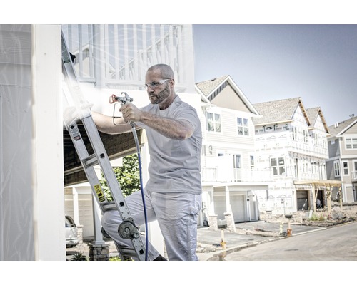 Muž na žebříku natírá dům stříkací pistolí