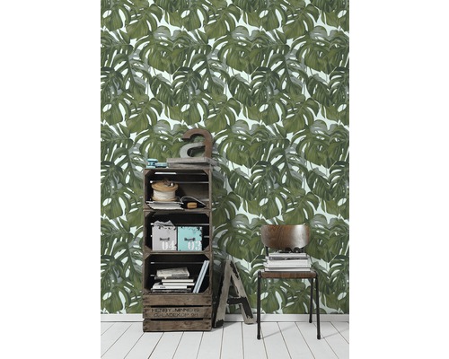 Obývací pokoj s tapetou s motivy listů monstery, regál z dřevěných beden a židle se stohom knih