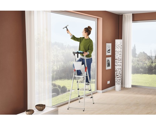 Žena myje okna stěrkou na okna ze schůdků v obývacím pokoji.