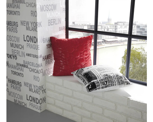 Dekorativní interiér s červeným a černo-bílým polštářem na parapetu před stěnou potištěnou názvy měst