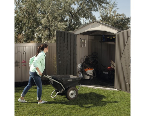 Žena tlačí kolečko do zahradního domku s otevřenými dveřmi, kde je umístěno zahradní nářadí.