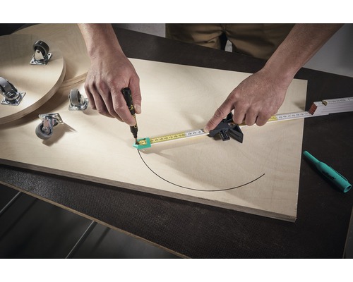 Osoba kreslí kružítkem a perem půlkruh na dřevěnou desku. Na desce jsou také nábytková kolečka.