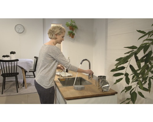 Světlá kuchyně se ženou u umyvadla, kohoutek, dřez a kuchyňské náčiní.