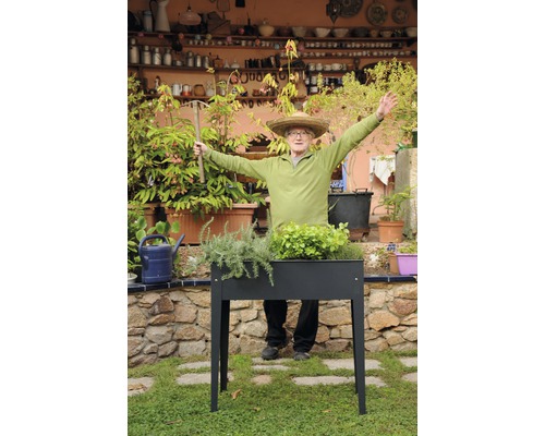 Muž se slaměným kloboukem stojí za vyvýšeným záhonem s rostlinami v zahradě.