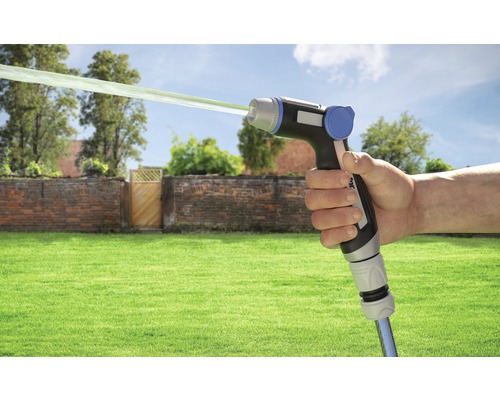 Ruka drží zahradní postřikovač s proudem vody před zahradní scenérií